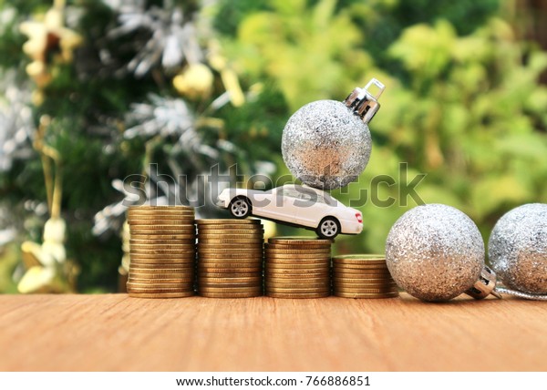 White miniature car carry silver glitter ball and\
drives on rolls ladder of gold coins money on wood table in blur\
natural tree