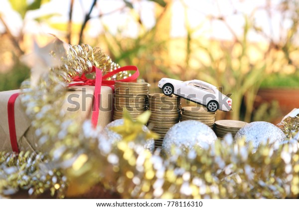 White miniature car drives on roll ladder of gold coin\
money lead to present box with Christmas glitter balls decoration\
and stars ribbons in blur natural tree in bright sunlight          \
        