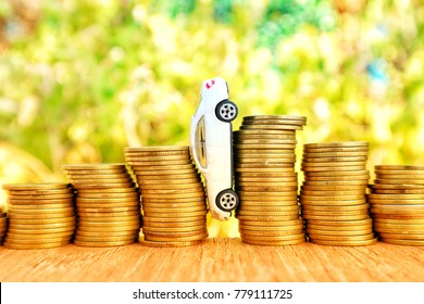 White Miniature Car Falls Between Roll Unstable Ladder Step Of Gold Coin Money On Wood Table In Blur Natural Tree In Bright Light                       