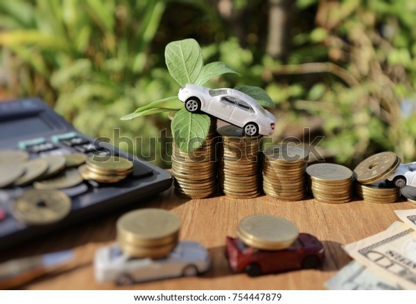 White miniature car with plant tree on\
top rolls ladder of gold coins and vehicles carry money, pile coins\
on calculator and banknote on wood table           \
