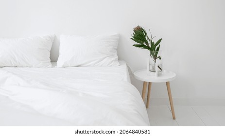 White Minimalist Apartment With Modern Interior Design. Banner View Of Comfort Bedroom With Beautiful Flower On Night Stand In Bright Morning Room. Soft And Clean Bedding On Bed. Hotel Service Concept