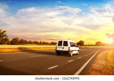White minivan driving fast on a countryside asphalt road with trees against an evening sky with a sunset