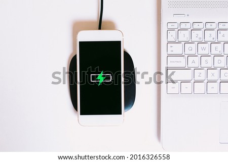 White mobile smart phone with the logo of a battery with green lightning on the screen, charging on a charger base without cable next to the laptop on a white work table