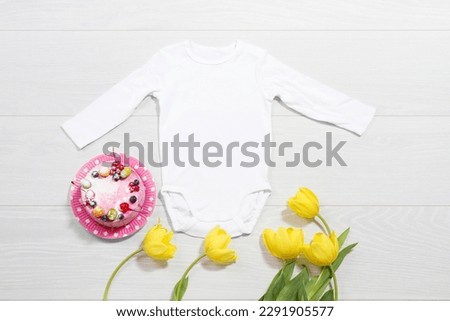 White mockup baby newborn shirt. Wooden background, yellow tulips, happy birthday pink cake. Blank template jumpsuit bodysuit front top view. Baby clothing. Bouquet of flowers