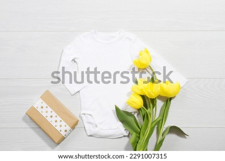 White mockup newborn shirt. Wooden background, yellow tulips, happy birthday gift box. Blank template jumpsuit bodysuit front top view. Baby clothing. Bouquet of flowers