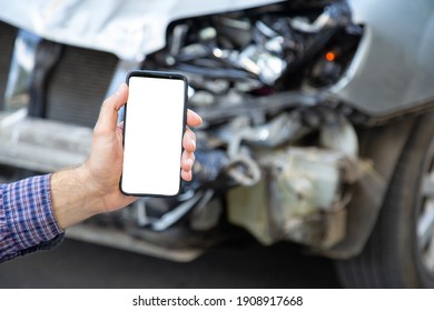 White Mockup Screen Mobile Phone In Male Hands After Car Accident. Mock Up For Insurance Service, Driving School, Tow Truck Call Or Web App Above Car Accident. Smartphone In Front Of Wrecked Car