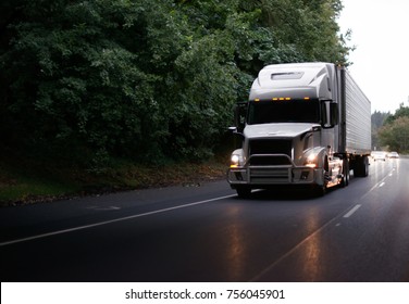 White modern big rig semi truck with grille guard and turn on headlights and reefer semi trailer running on evening road with green trees and light reflection on the road surface