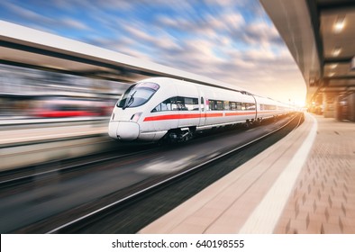 White Modern High Speed Train In Motion On Railway Station At Sunset. Train On Railroad Track With Motion Blur Effect In Europe In Evening. Railway Platform. Industrial Landscape. Railway Tourism