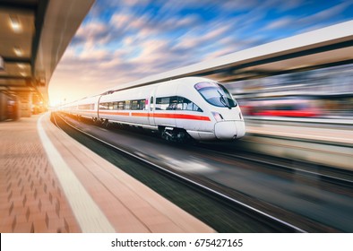 White modern high speed train in motion on railway station at sunset. Train on railroad track with motion blur effect in Europe in evening. Railway platform. Industrial landscape. Railway tourism