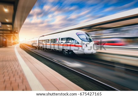 White modern high speed train in motion on railway station at sunset. Train on railroad track with motion blur effect in Europe in evening. Railway platform. Industrial landscape. Railway tourism