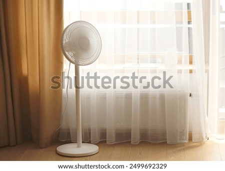 A white, modern pedestal fan is positioned near a window with curtains. The scene is well-lit with natural light coming through the window, creating a calm and airy ambiance.