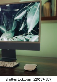 White Mouse And Keyboard In Front Of Large Computer Screen On A Wood Table With Bookshelf And Soft Green Light In The Background 