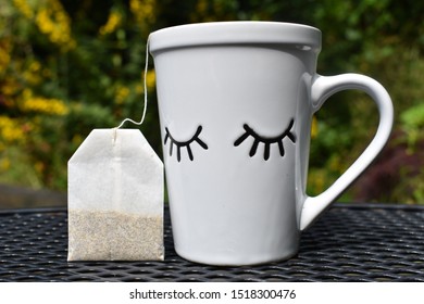 White Mug With Closed Eye Detail And Teabag On A Table