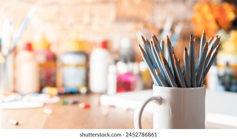 White mug with colored pencils in the foreground on a wooden table with art supplies in the background out of focus - Powered by Shutterstock