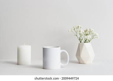 White mug mockup with beige vase, candle and gypsophila branch on a table