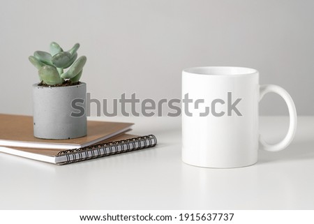 White mug mockup on table in minimalist interior, business concept, succulent and notepads. Template, layout for your design, advertising, logo with copy space. Cup on light gray background.