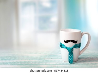 White Mug On Wooden Table With Mustache And Tie Indoors Blue Empty Space Background.Fathers Day Concept.Masculine Wallpaper. 