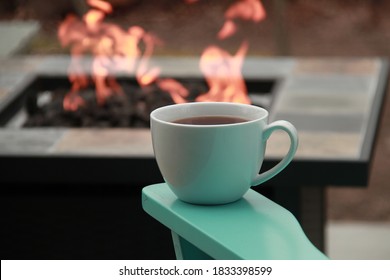 White Mug Of Tea On The Arm Of A Teal Blue Chair With A Fire In A Fire Pit In The Background On A Stone Patio Outside