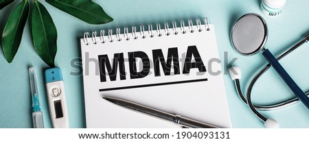In a white notebook on a blue background, near a sheet of shefflers, a stethoscope, a syringe and an electronic thermometer, the word MDMA is written. Medical concept