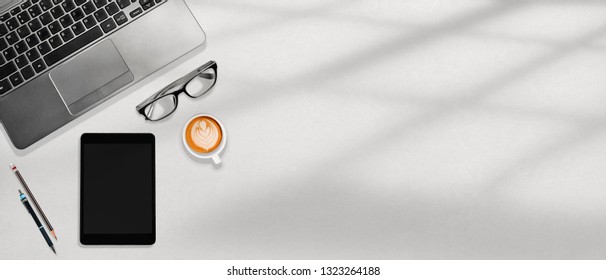 White Office Desktop Workspace, Work Place Flatlay With Part Of Laptop, Tablet, Eye Glasses And A Cup Of Coffee With Froth Art. Natural Window Light And Shadow On The Table. Copy Space.