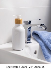 White one pump bottle with bamboo tap on basin near water tap and folded blue towel in bathroom close up, cosmetic mockup.  Minimal composition for brand packaging