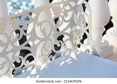 White openwork wrought iron bench on the sea embankment. Close-up, diagonal composition. A beautiful summer picture on the theme of relaxing in a park by the sea during the holiday season.