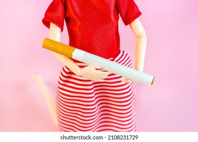 White And Orange Cigarette In Woman Doll Hands On Light Pink Background. Children Smoking And Youth Tobacco Use Addiction Concept