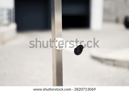 White outdoor CCTV security camera mounted on metal pole. Surveillance camera monitoring parking lot entrance and property territory.