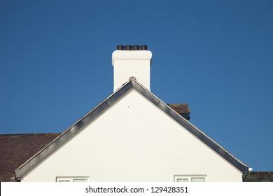 A white painted gable end with plastic barge boards a chimney and pots with a slate roof against a blue sky.