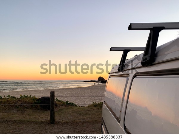 A white panel\
van with a roof rack parked up in  a beach side car park. The\
rising sun casts colours onto the horizon and they reflect off the\
side of the vans white wall