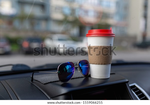 White\
paper coffee Cup and sunglasses on the dashboard of the car. Paper\
Cup with hot tea and glasses on the dashboard of the SUV close up\
against the background of a blurred Parking\
lot
