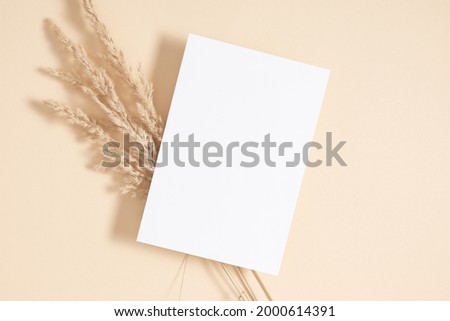White paper empty blank, dried grass decoration on beige background. Invitation card mockup on beige table. Flat lay, top view, copy space, mockup