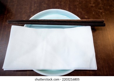 White paper serviettes with detailed embossing on a plate. A pair of wooden chopsticks located above as neat table arrangement in a Chinese restaurant. - Shutterstock ID 2203216805