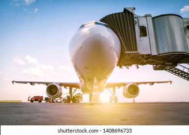 A white passenger airplane stands at the boarding bridge and is loaded with baggage in the rays of the morning sun
