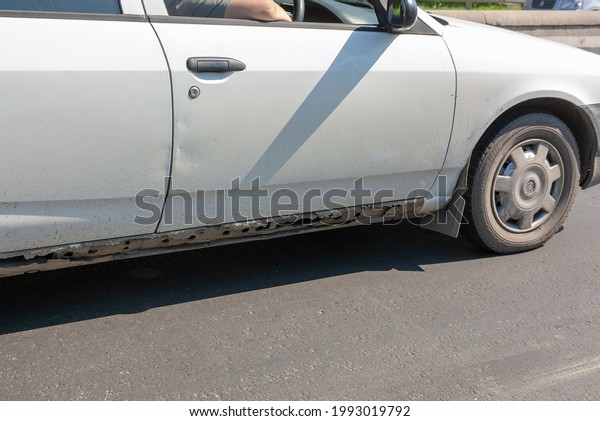A white passenger car is moving along the road.\
On the lower part of the car body, traces of through-corrosion of\
the metal with the destruction of the threshold of the car are\
clearly visible.