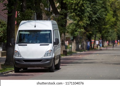 White Passenger Medium Size Commercial German Luxury Minibus Van Parked On City Street With Blurred Silhouettes Of Pedestrians And Moving Car Under Green Trees.