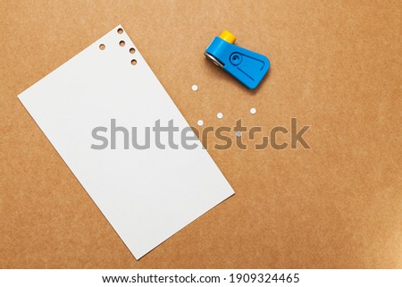 White perforated paper with a hole puncher placed on a cardboard surface. The hole circles are next to the sheet along with the blue hole punch and yellow button.