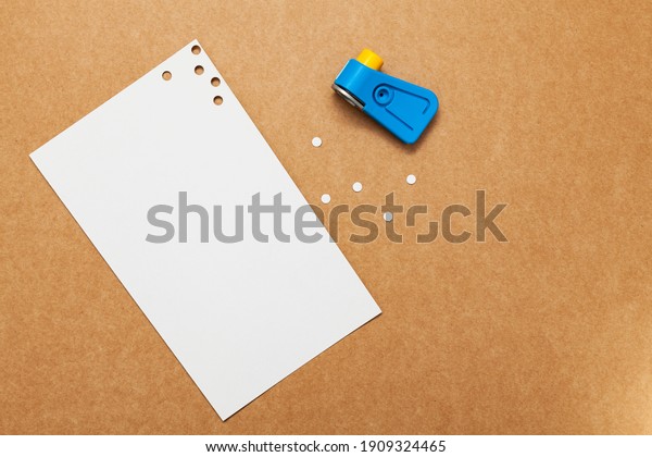 White perforated paper with a\
hole puncher placed on a cardboard surface. The hole circles are\
next to the sheet along with the blue hole punch and yellow\
button.