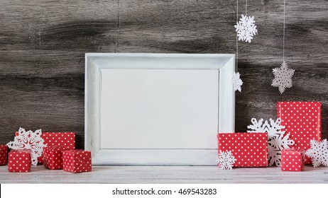 White Photo Frame With Christmas Decoration On The Wooden Table. Red Boxes And Snowflakes On The Grey Background. Frame For Quotes. Christmas Postcard.