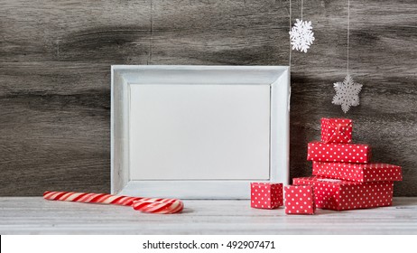 White Photo Frame With Christmas Decoration On The Wooden Table. Red Boxes And Snowflakes On The Grey Background. Frame For Quotes. Christmas Postcard.