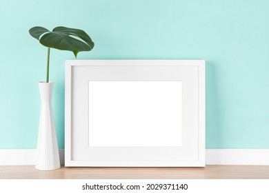 White picture frame with matte and monstera deliciosa leaf in vase in front of light turquoise wall. Elegant poster mockup, blank image area isolated with clipping path.