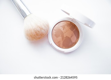 A white pillbox with powder and bronzer lies on a white background, next to it lies a brush for blush. High quality photo