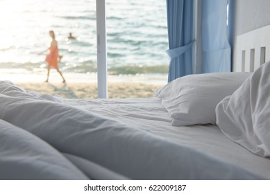 White pillows and bedding sheet in clean hotel bedroom with woman walking on the beach, sea view background, happy summer holiday at luxury resort