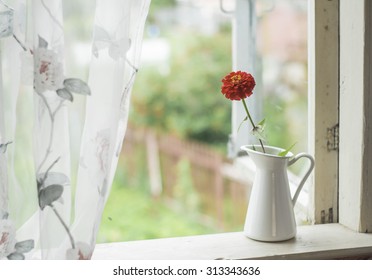 White pitcher stands at the window. Red flower in a white pitcher. Window in a country house.