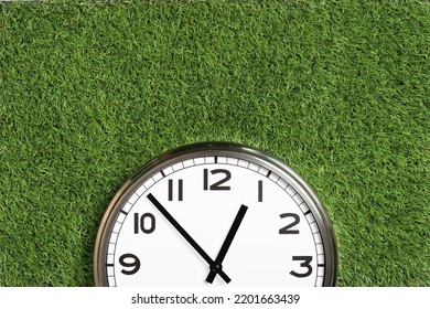 White Plain Analogue Wall Clock On Green Turf Grass Background. Five To One O'clock. Copy Space, Game Time Management Concept. Opening Or Closing Hours. Ecological Sustainable Business Banner