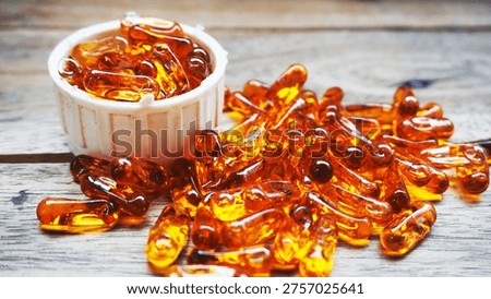 White plastic container filled with translucent amber softgel capsules, with additional capsules scattered on a wooden surface. The glossy texture and vibrant color highlight dietary supplements or medications, often associated with fish oil, vitamin E, or oil-based nutrients.                               