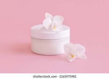 White plastic cream jar near white orchid flowers on light pink, close up, mockup. Skincare beauty product, cream or lotion. Exotic natural cosmetics, pastel minimal composition