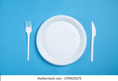 White Plastic Plate, Fork And Knife On Blue Background.