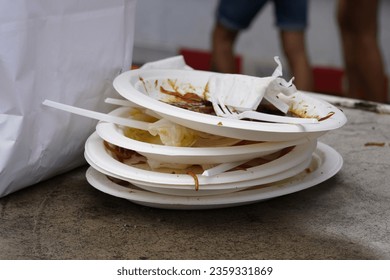 White plastic plates are stacked on one another. There are rests of the food on the  plates as well as some pieces of white plastic cutlery. On the defocused background there are passers by. 