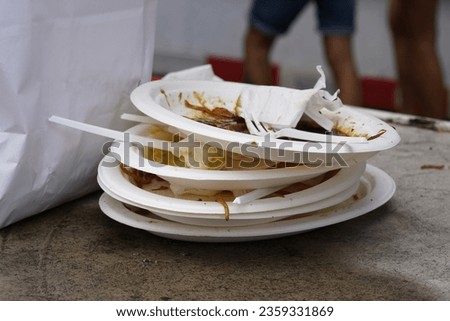 White plastic plates are stacked on one another. There are rests of the food on the  plates as well as some pieces of white plastic cutlery. On the defocused background there are passers by. 